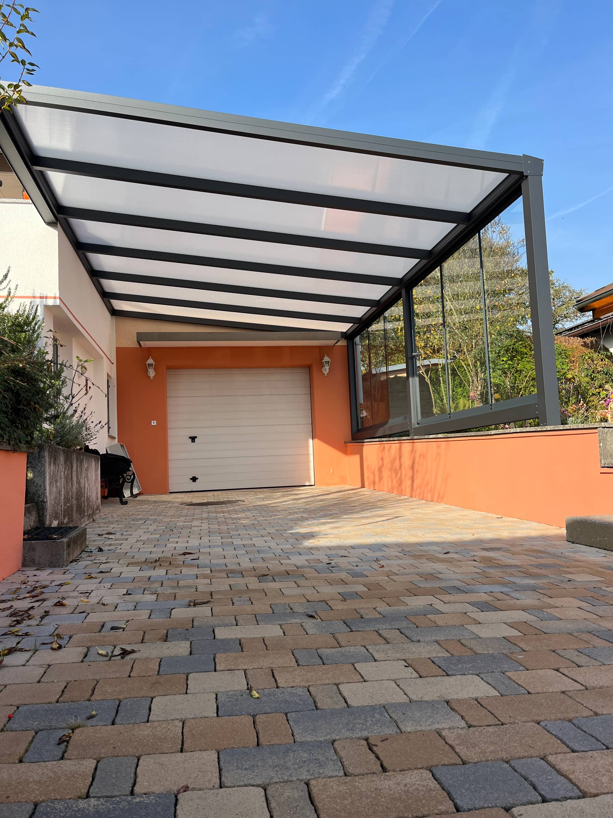 Carport aus Aluminium in Anthrazit mit lichtdurchlässiger Eindeckung, an das Wohnhaus angebaut über gepflasterter Zufahrt