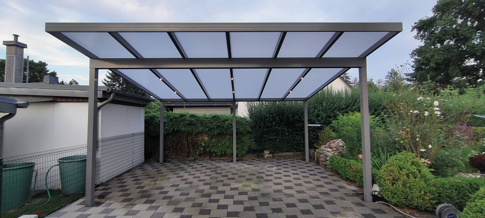 Carport aus Aluminium mit lichtdurchlässiger Dachfläche über gepflastertem Stellplatz im Garten