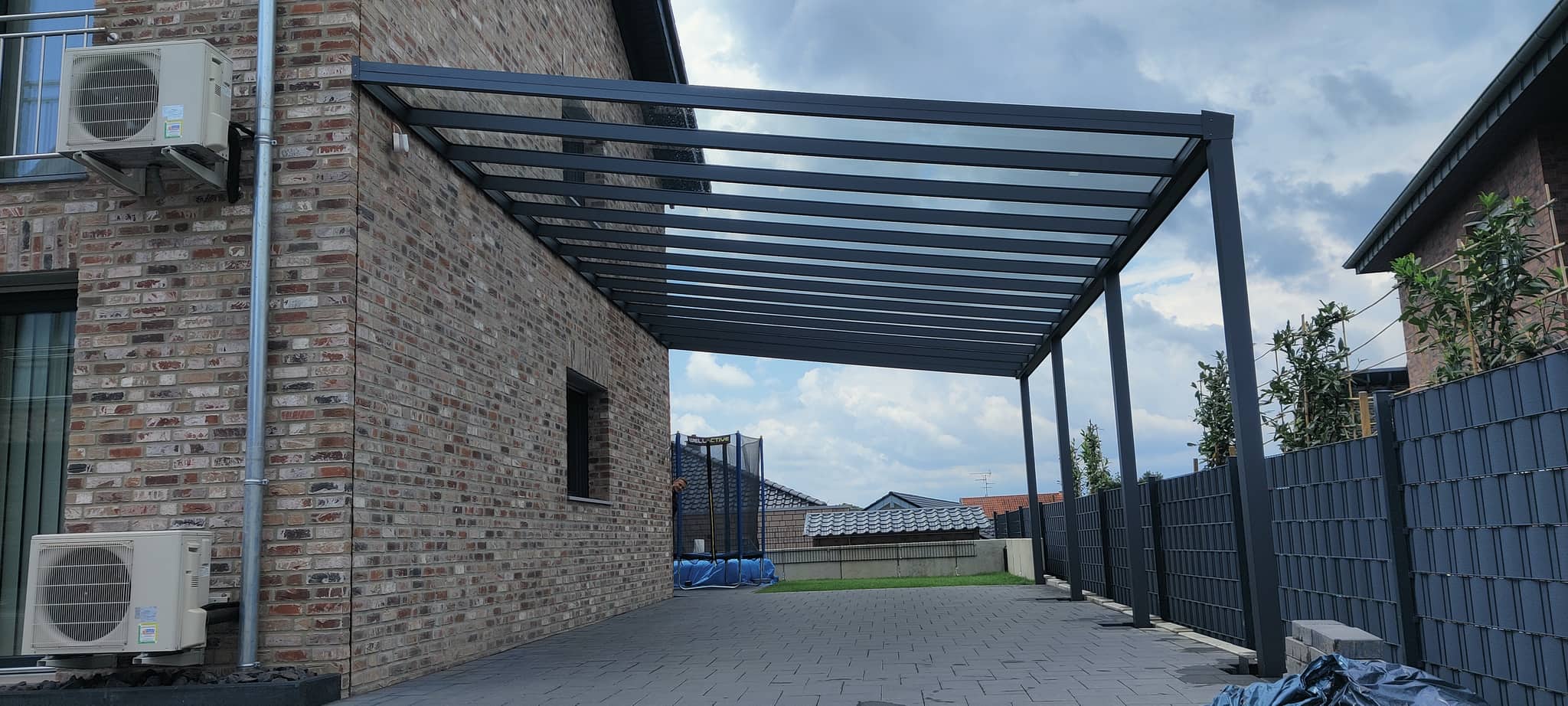 Pultdach-Carport aus Aluminium in Anthrazit mit lichtdurchlässiger Eindeckung zwischen zwei Klinkerhäusern, blauer Himmel