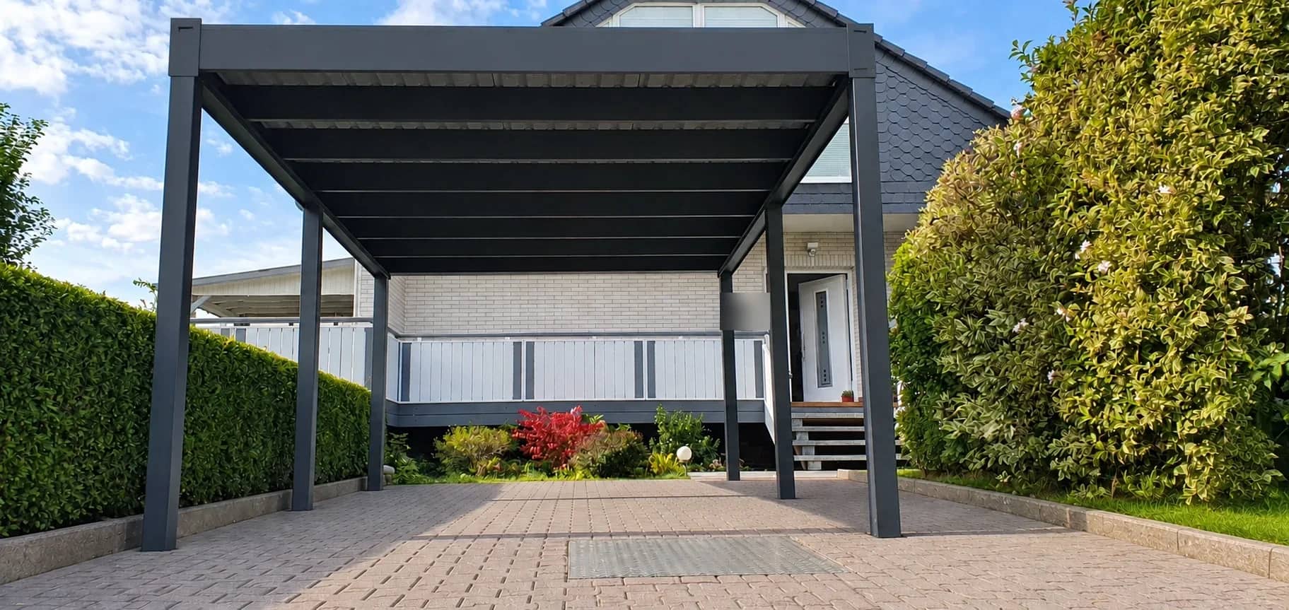 Flachdach-Carport aus Aluminium in Anthrazit über einer gepflasterten Einfahrt vor einem Wohnhaus, seitlich Hecken