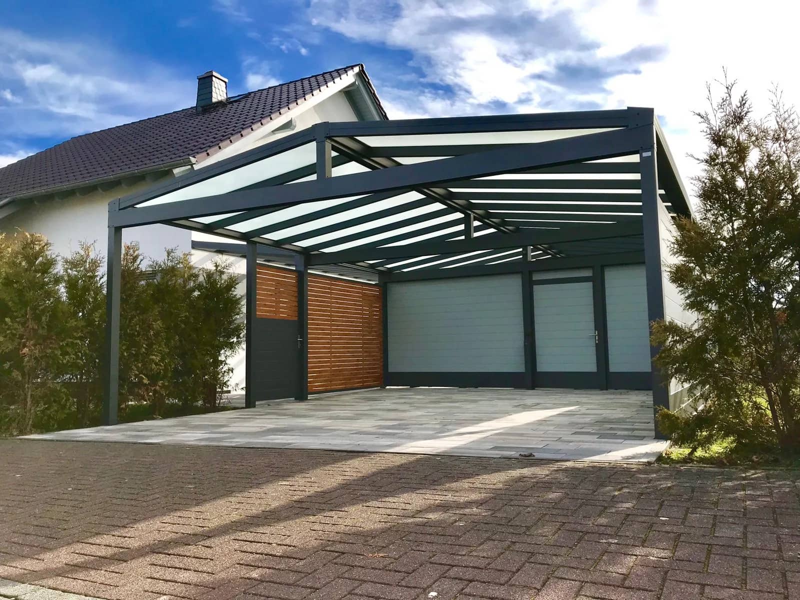 Carport aus Aluminium in Anthrazit mit lichtdurchlässiger Eindeckung vor einer Garage, seitlich ein Element in Holzoptik