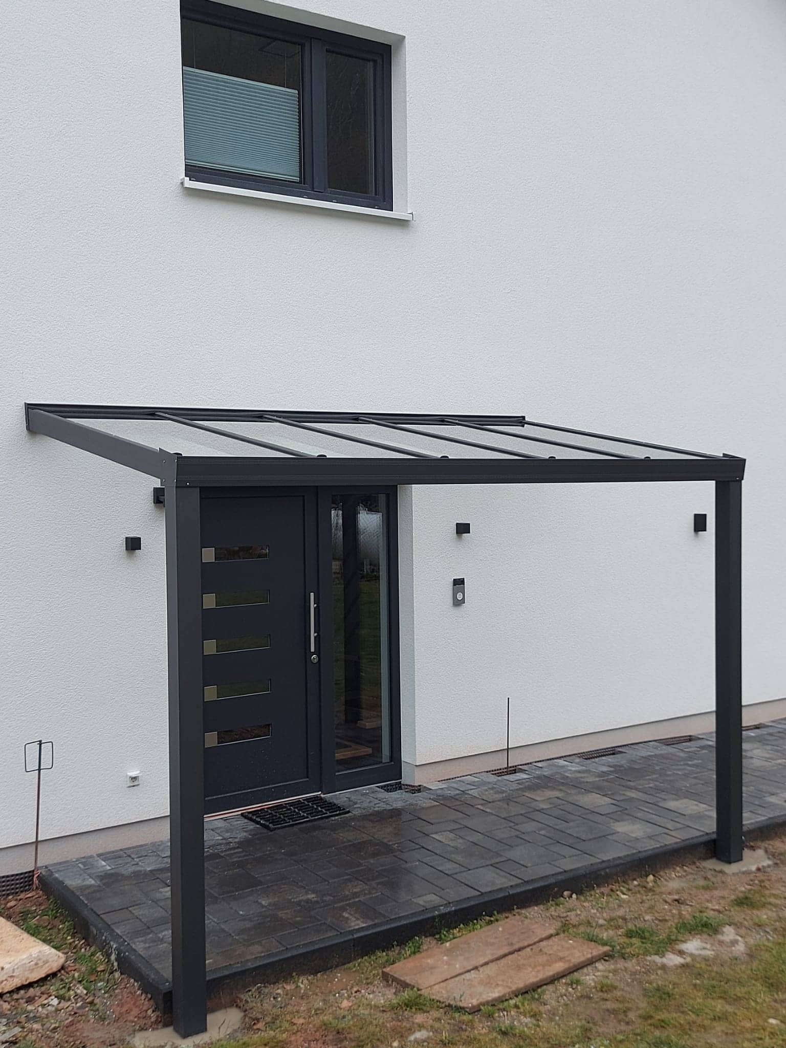 Eingangsüberdachung aus Aluminium in Anthrazit mit lichtdurchlässiger Eindeckung über der Haustür eines hellen Wohnhauses