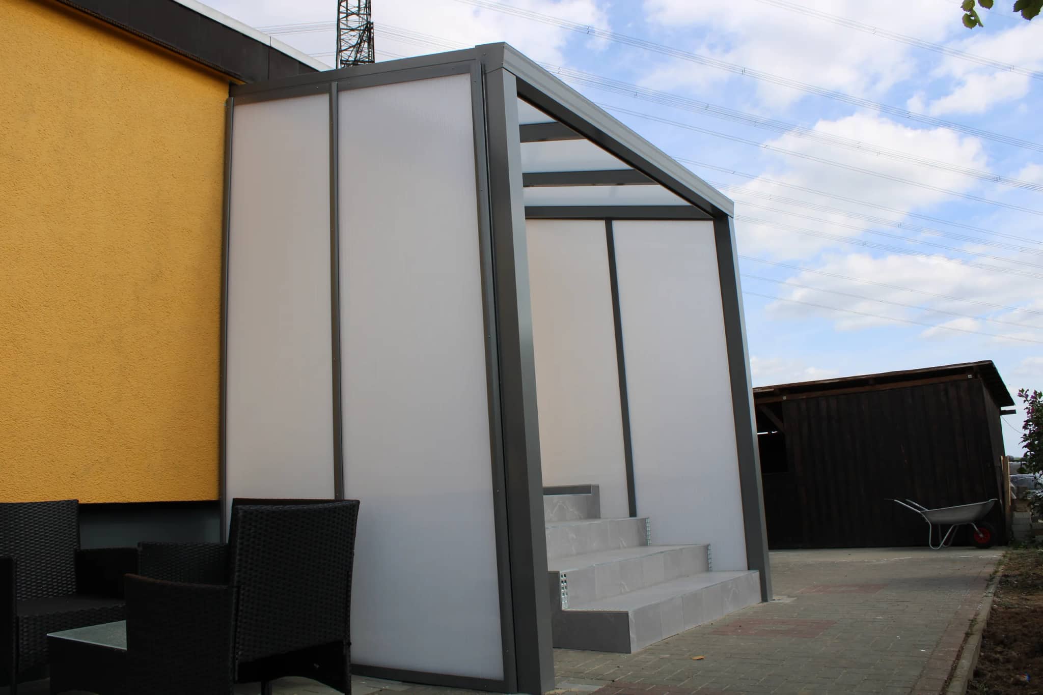 Eingangsüberdachung aus Aluminium in Anthrazit mit lichtdurchlässigen Seitenwänden über der Außentreppe eines gelben Wohnhauses