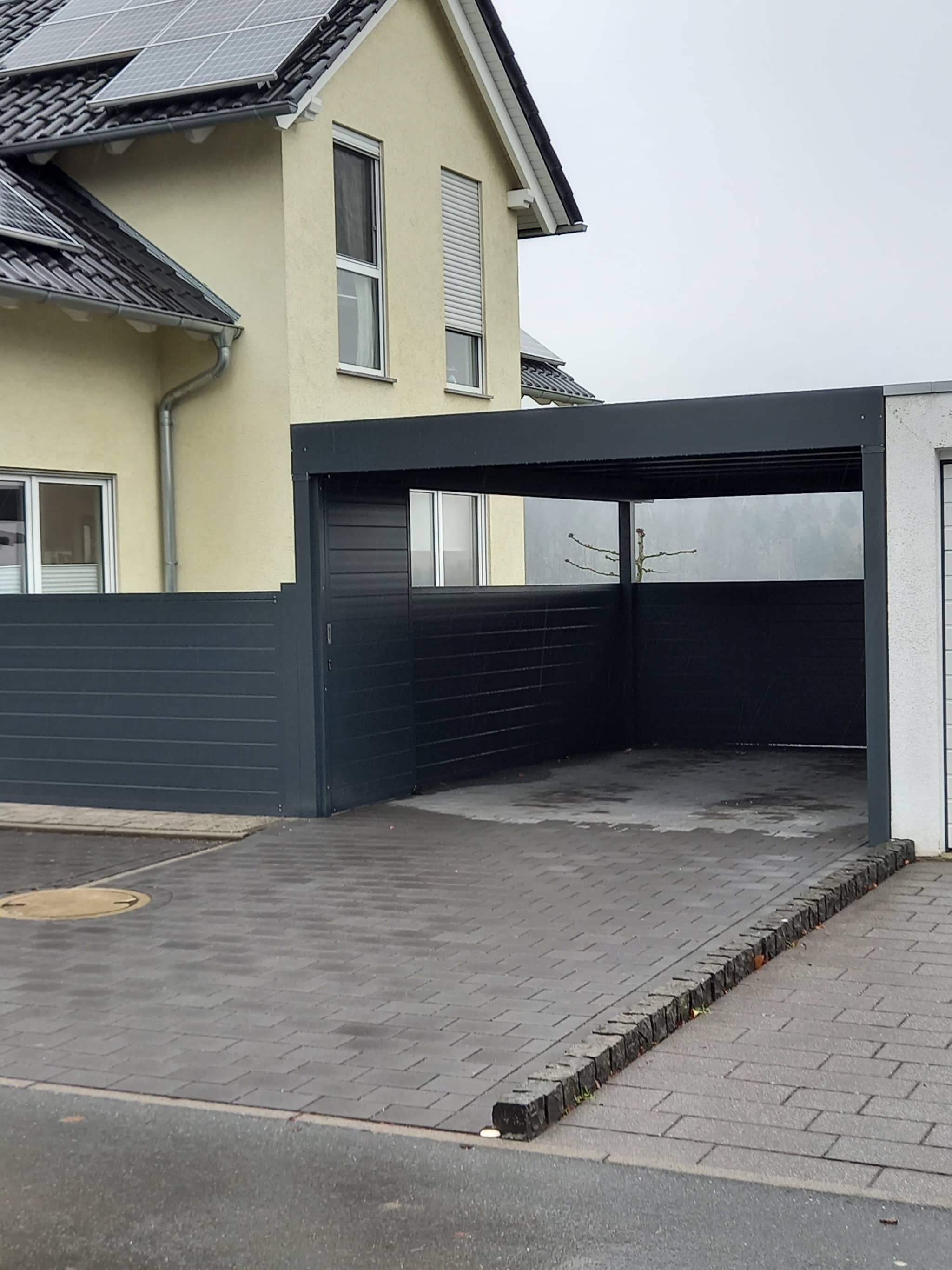 Carport in Anthrazit direkt am Wohnhaus, mit geschlossenen Seitenwänden auf gepflasterter Einfahrt