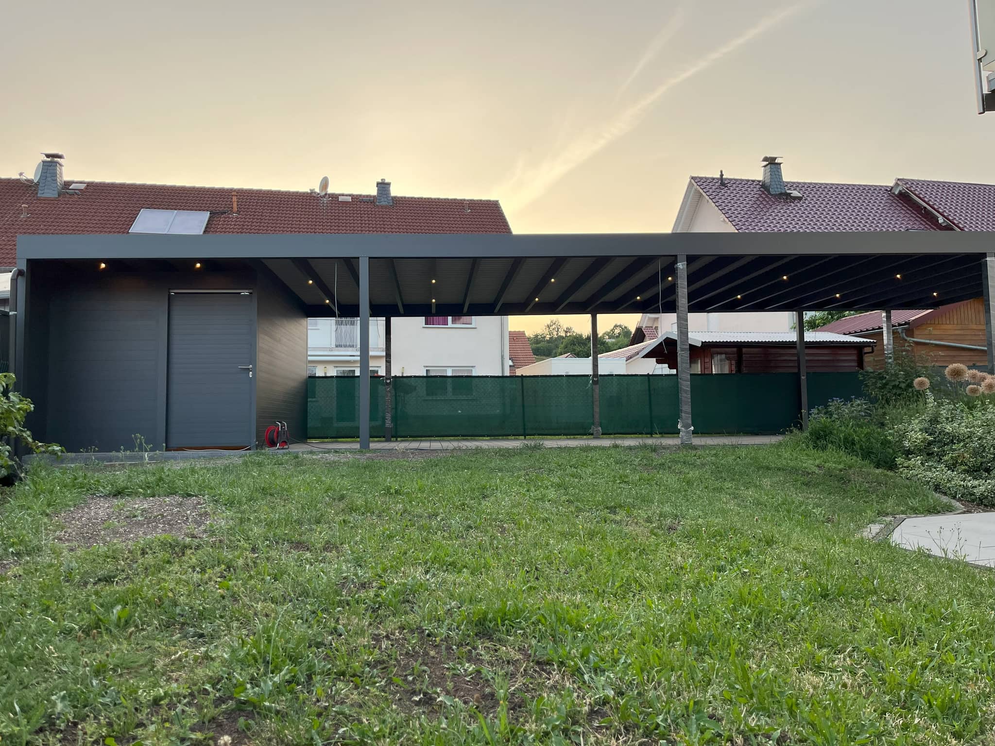 Flachdach-Carport aus Aluminium in Anthrazit mit geschlossenem Abstellraum und Einbauleuchten in der Dachuntersicht, davor Rasen, Abendhimmel
