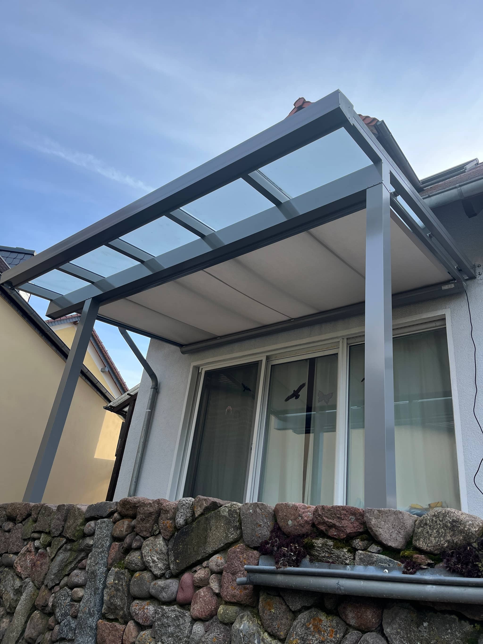 Unterdachmarkise mit hellem Tuch unter einem Aluminium-Terrassendach, darunter eine Bruchsteinmauer