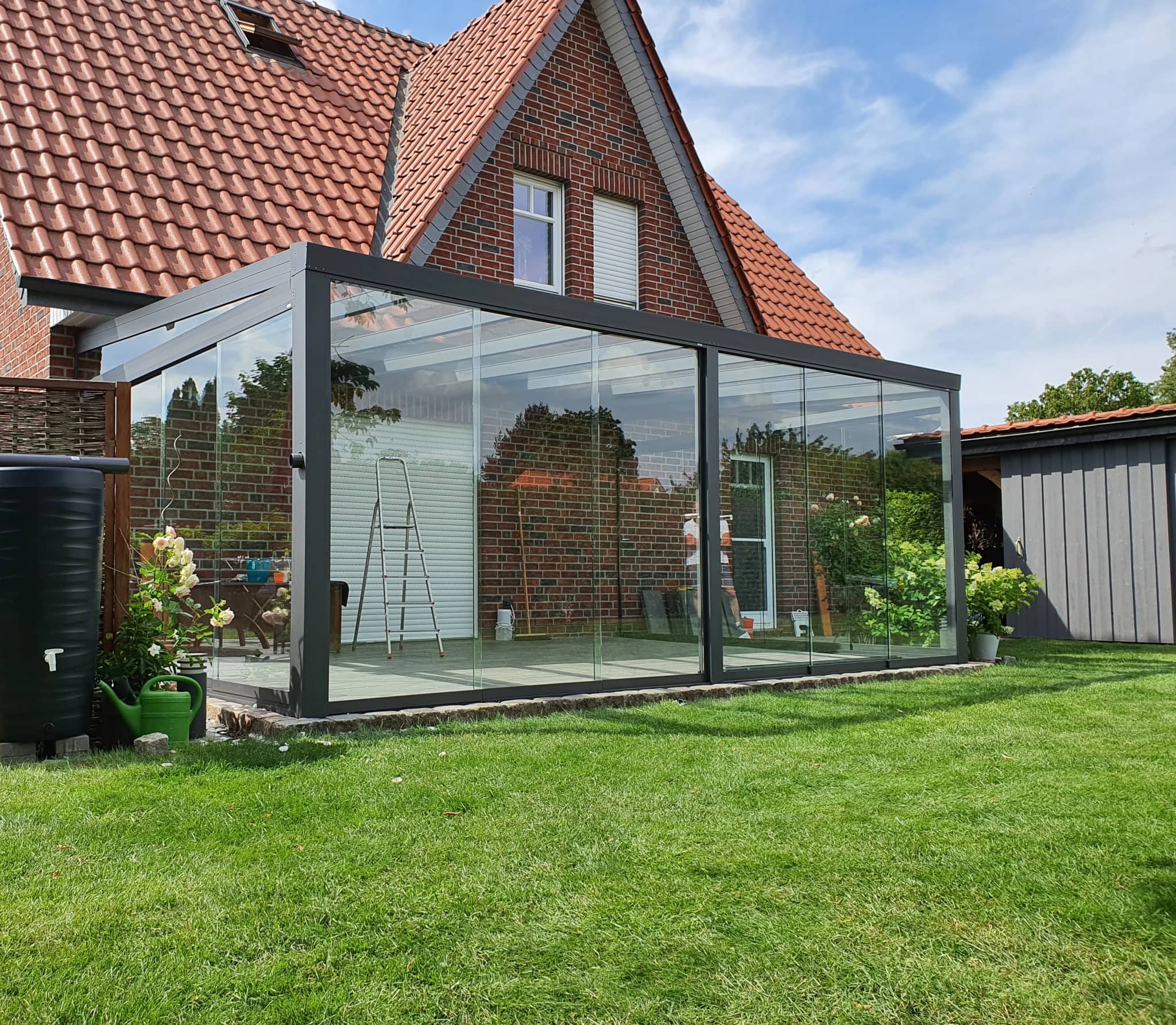 Sommergarten in Anthrazit mit rundum verglasten Schiebewänden als Anbau an ein Backsteinhaus mit Ziegeldach