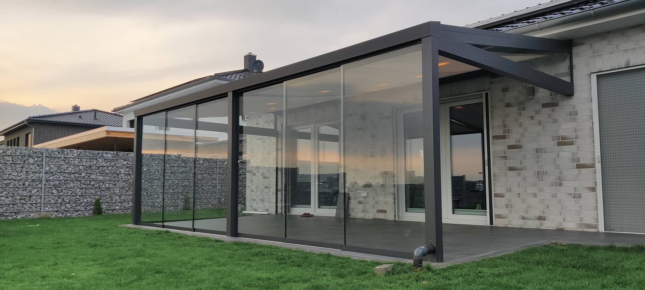 Terrassenüberdachung aus Aluminium mit umlaufenden Glas-Schiebewänden in der Abendstimmung, daneben ein Gabionenzaun