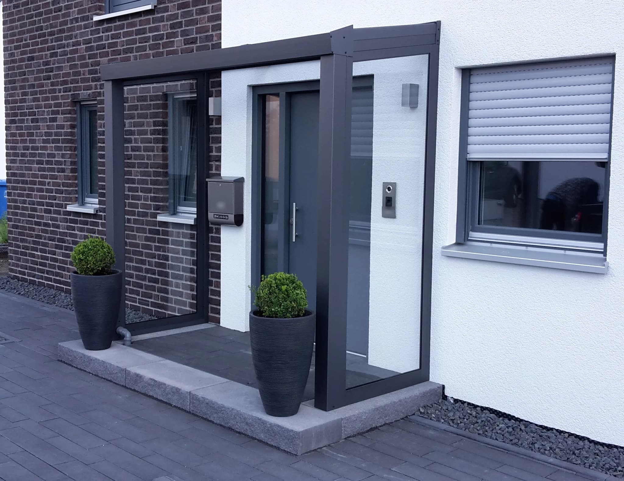 Eingangsüberdachung aus anthrazitfarbenem Aluminium mit gläsernem Seitenteil, wettergeschützter Hauseingang an heller Putzfassade mit Klinker