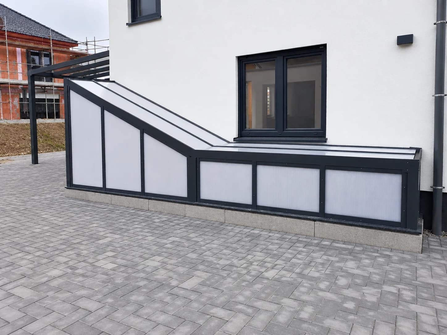 Eingangsüberdachung aus Aluminium in Anthrazit über einem Treppenabgang mit lichtdurchlässigen Seitenelementen an einem weißen Wohnhaus