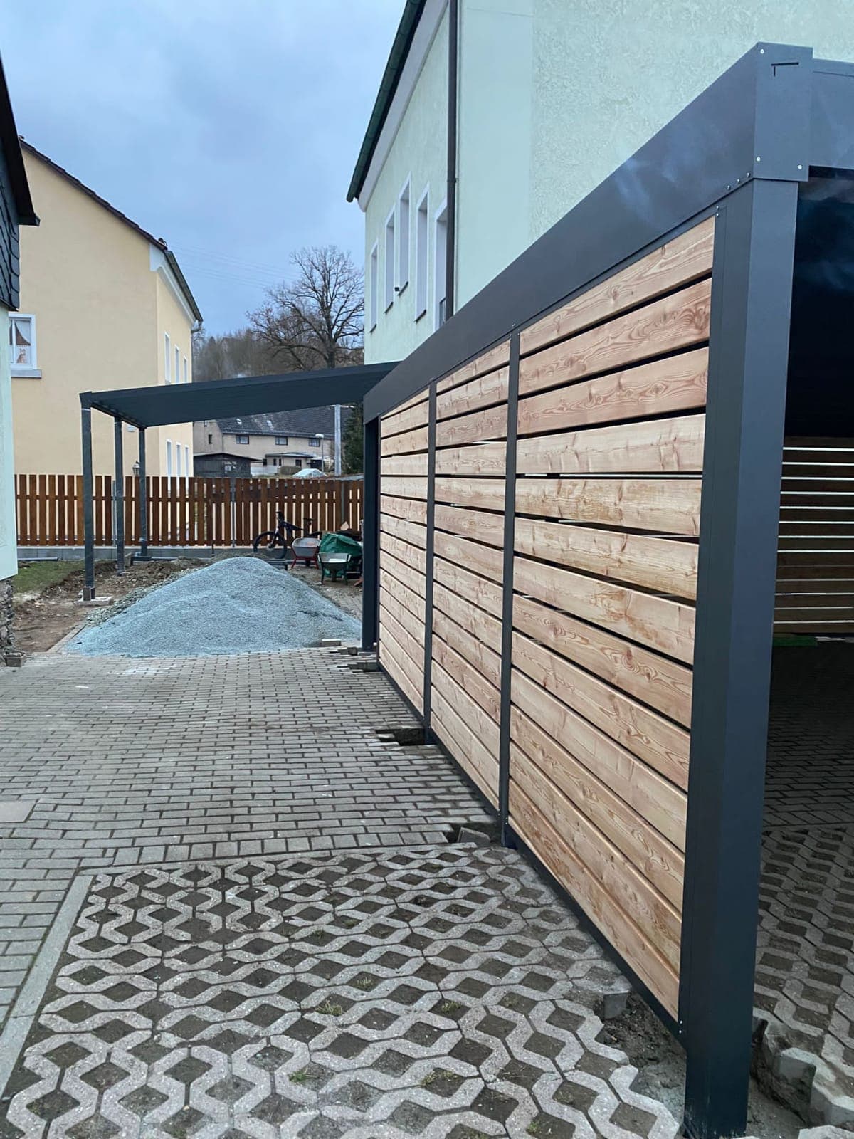 Carport aus Aluminium in Anthrazit mit seitlichem Sichtschutz in Holzoptik über einer Einfahrt mit Rasengittersteinen